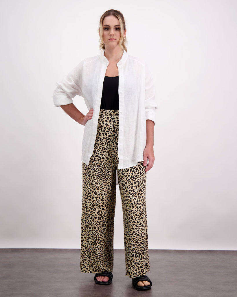 A standing female with caucasian skin and sandly coloured curly hair is wearing a black camisole under a white open linen shirt. She is wearing leopard print wide leg pants and black leather slippers. Christina Stephens Australian Adaptive Clothing.