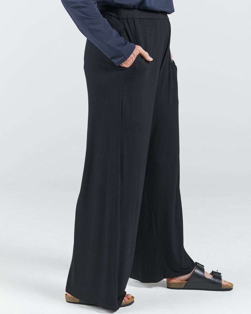 Image is of a female wearing a pair of black bamboo wide leg pants and black leather slippers. She is facing side-on. Her hands are in her pockets. Christina Stephens Australian Adaptive Clothing.