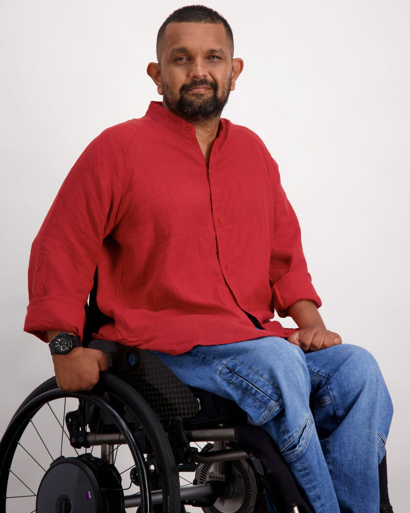 A male in a wheelchair with dark skin tone and dark hair is wearing a red linen shirt and a pair of blue denim adaptive jeans. Christina Stephens Australian Adaptive Clothing.  