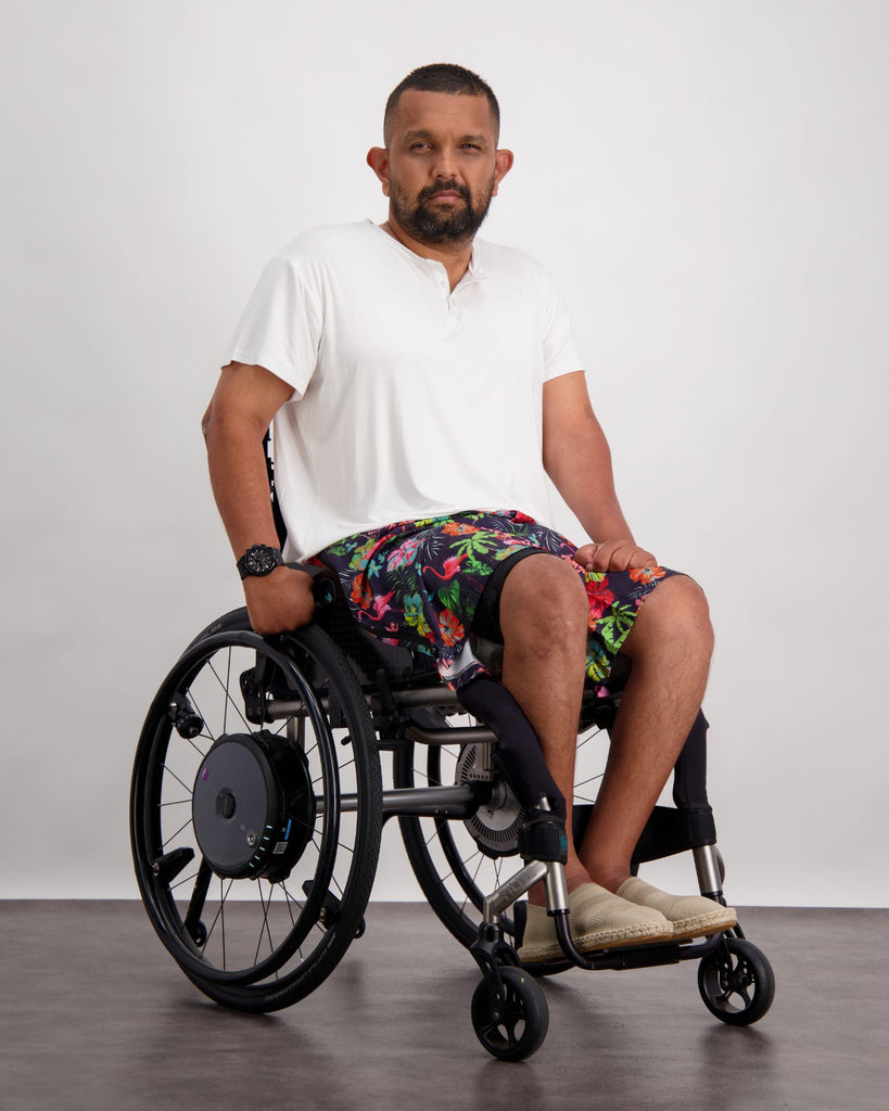 Male with dark skin and dark hair is sitting in a wheelchair wearing a white t-shirt, black and flamingo print board shorts and sand coloured loafer shoes. Christina Stephens Adaptive Clothing Australia. 