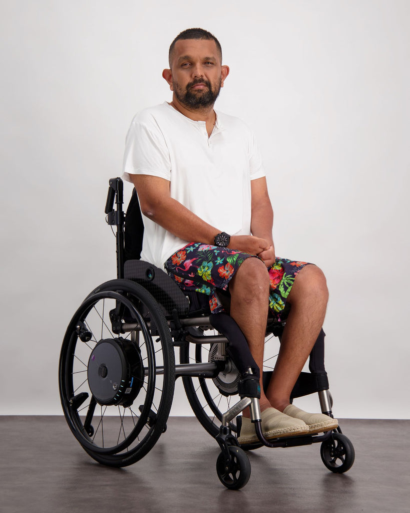 Male with dark skin and dark hair is sitting in a wheelchair wearing a white t-shirt, black and flamingo print board shorts and sand coloured loafer shoes. Christina Stephens Adaptive Clothing Australia. 