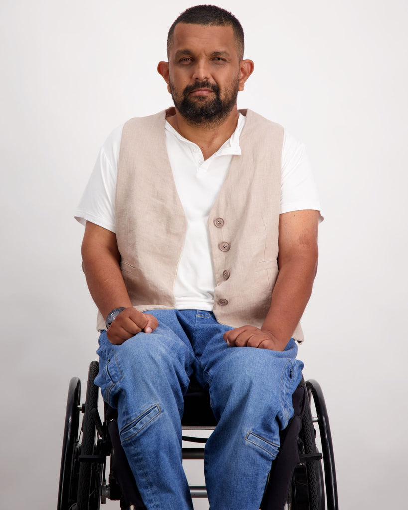 Male with dark skin and dark hair is sitting in a wheelchair wearing a white t-shirt, sand coloured linen vest with buttons and blue jeans. Christina Stephens Adaptive Clothing Australia. 