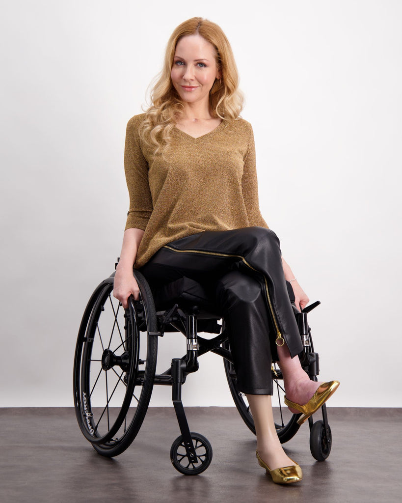 A blonde women sitting in a wheelchair wearing a gold glitter top and black pleather pants with gold zips up the sides of the legs. She is wearing gold shoes. Christina Stephens Adaptive Clothing Australia. 