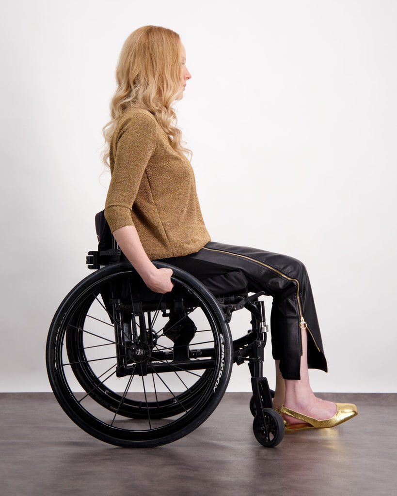 A blonde women sitting in a wheelchair (facing side on) wearing a gold glitter top with 3/4 length sleeves and black pleather pants with gold zips up the sides of the legs. She is wearing gold shoes. Christina Stephens Adaptive Clothing Australia.