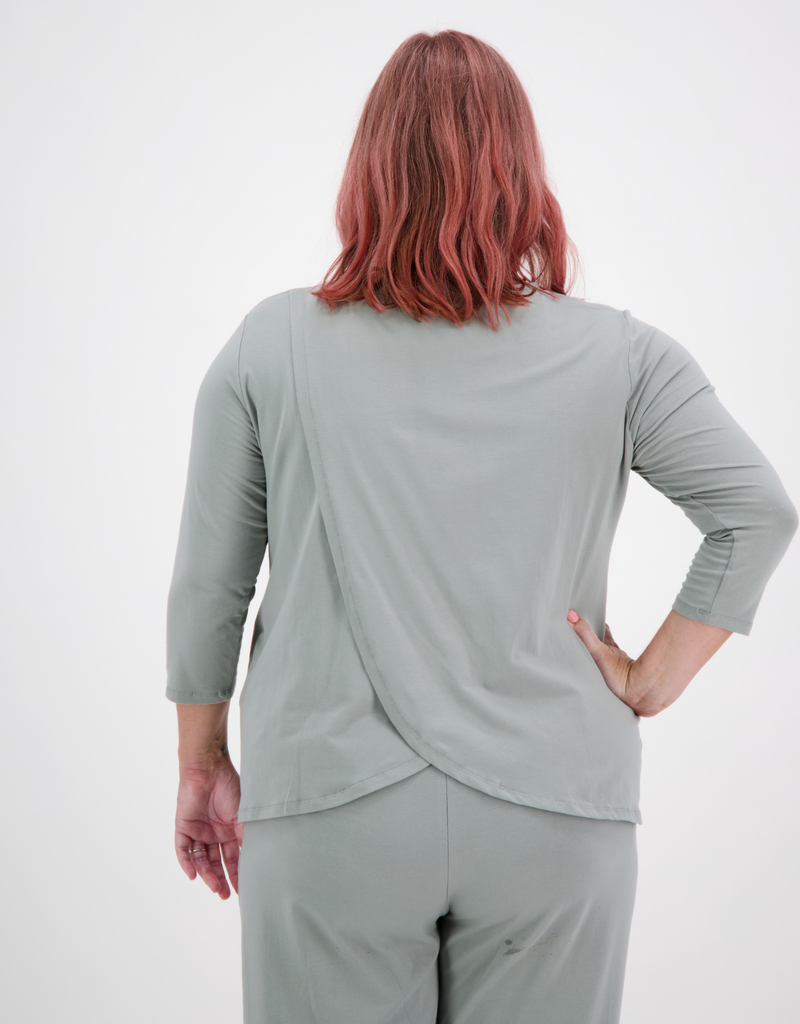 Woman with pink hair is wearing a sage green leaf back t-shirt with 3/4 length sleeves. He is facing to the back and paired the t-shirt with sage green trousers. 