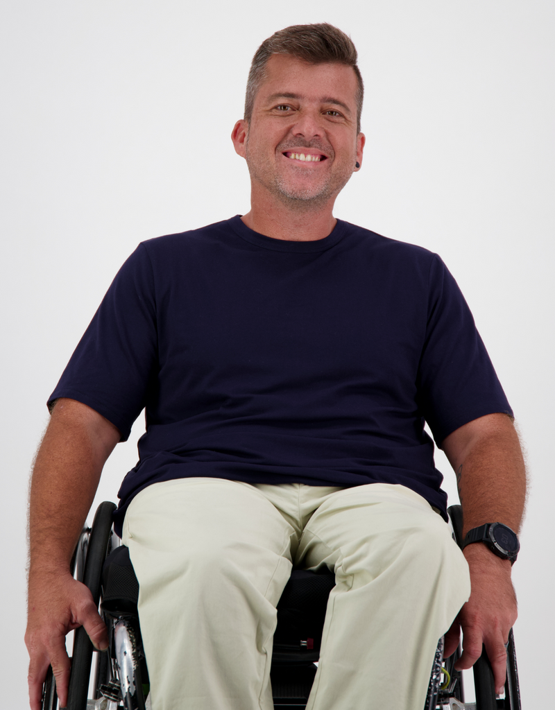 Man with light brown, short hair, is in a wheelchair wearing a navy t-shirt and cream cargo pants. Christina Stephens Australian Adaptive Clothing. 
